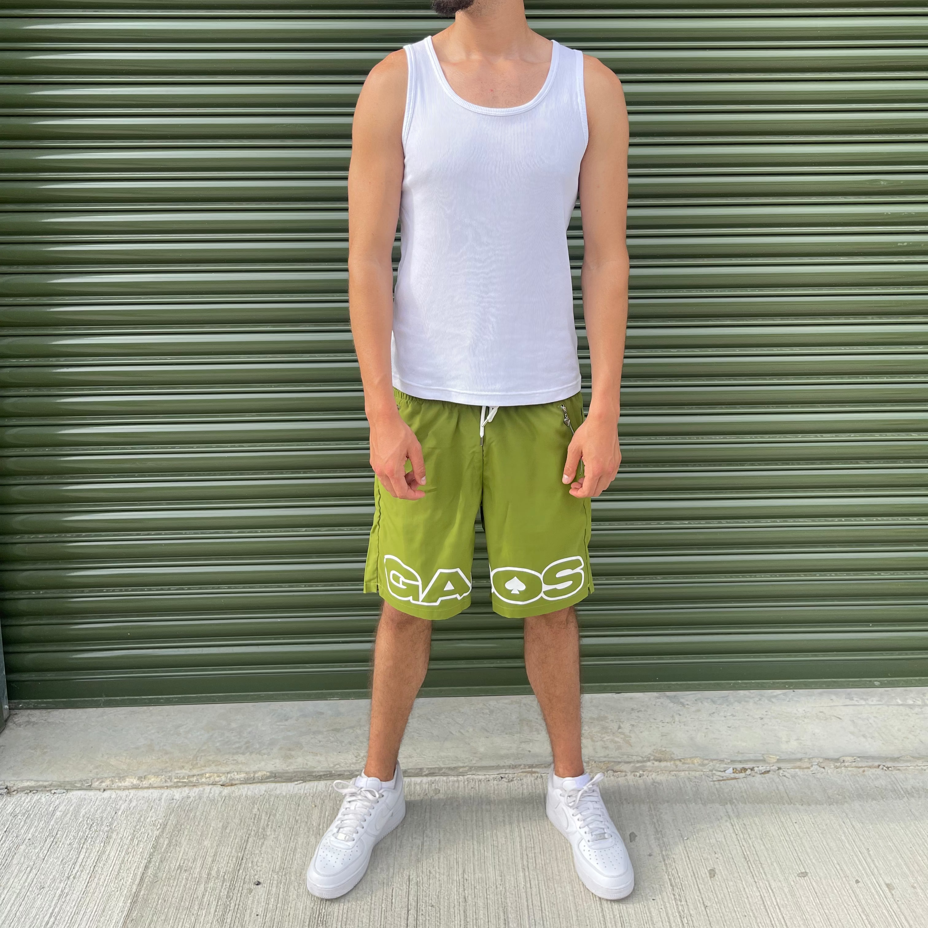 Man wearing a white tank top and gabos green shorts standing in front of a green shutter.
