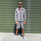 Model in grey Gabos London 3-piece tracksuit, pairing lightweight zip jacket and cargo-style shorts, seated casually in front of industrial building with sunglasses and white sneakers