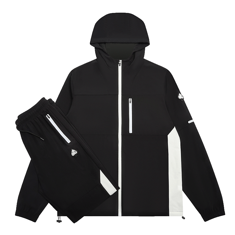 A black hooded jacket and matching black pants laid out on a white background. The jacket has a front zipper, a vertical chest zipper, a vertical white stripe on the side, and small white logo details.