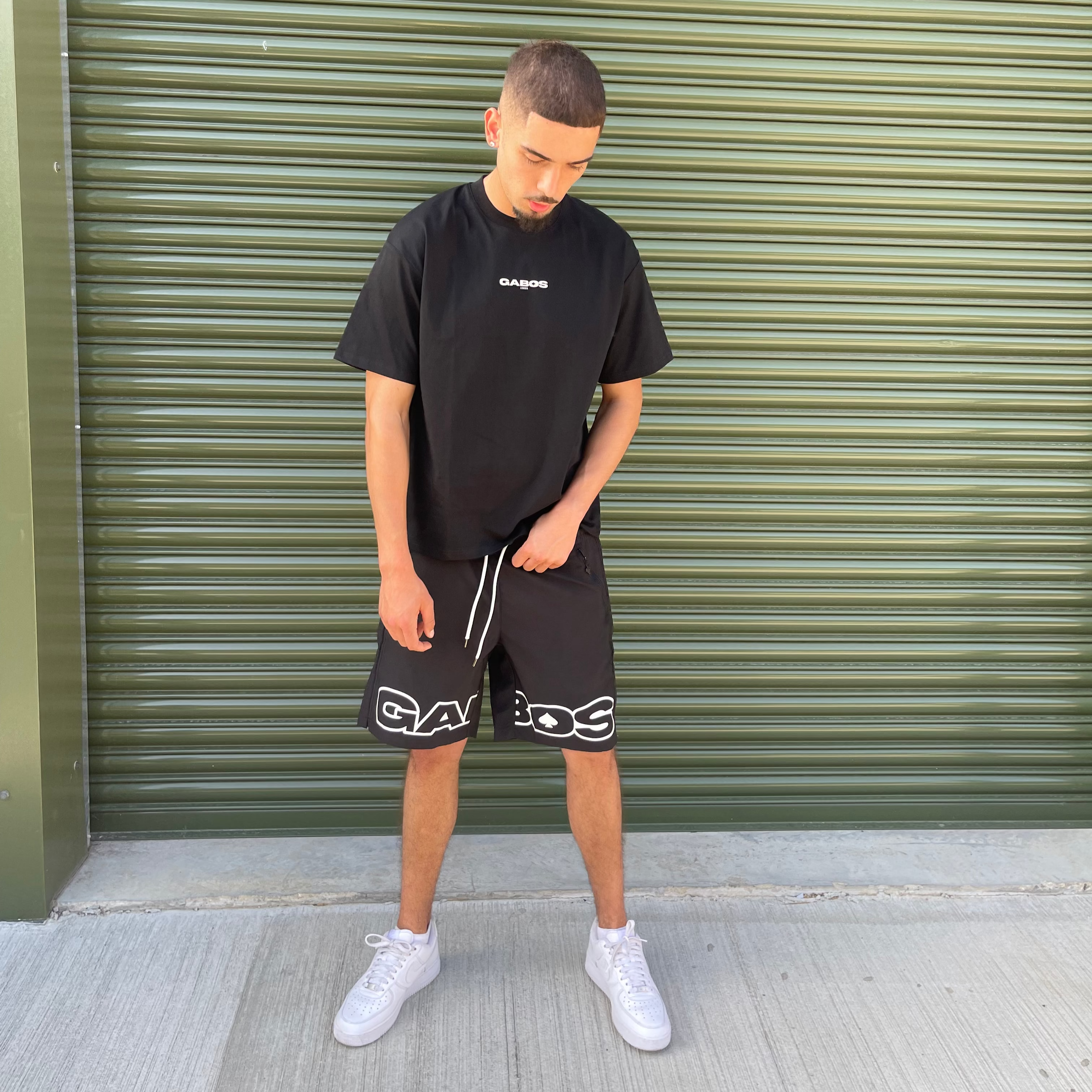 Person wearing a black t-shirt and shorts with visible gabos branding, standing in front of a green shutter.