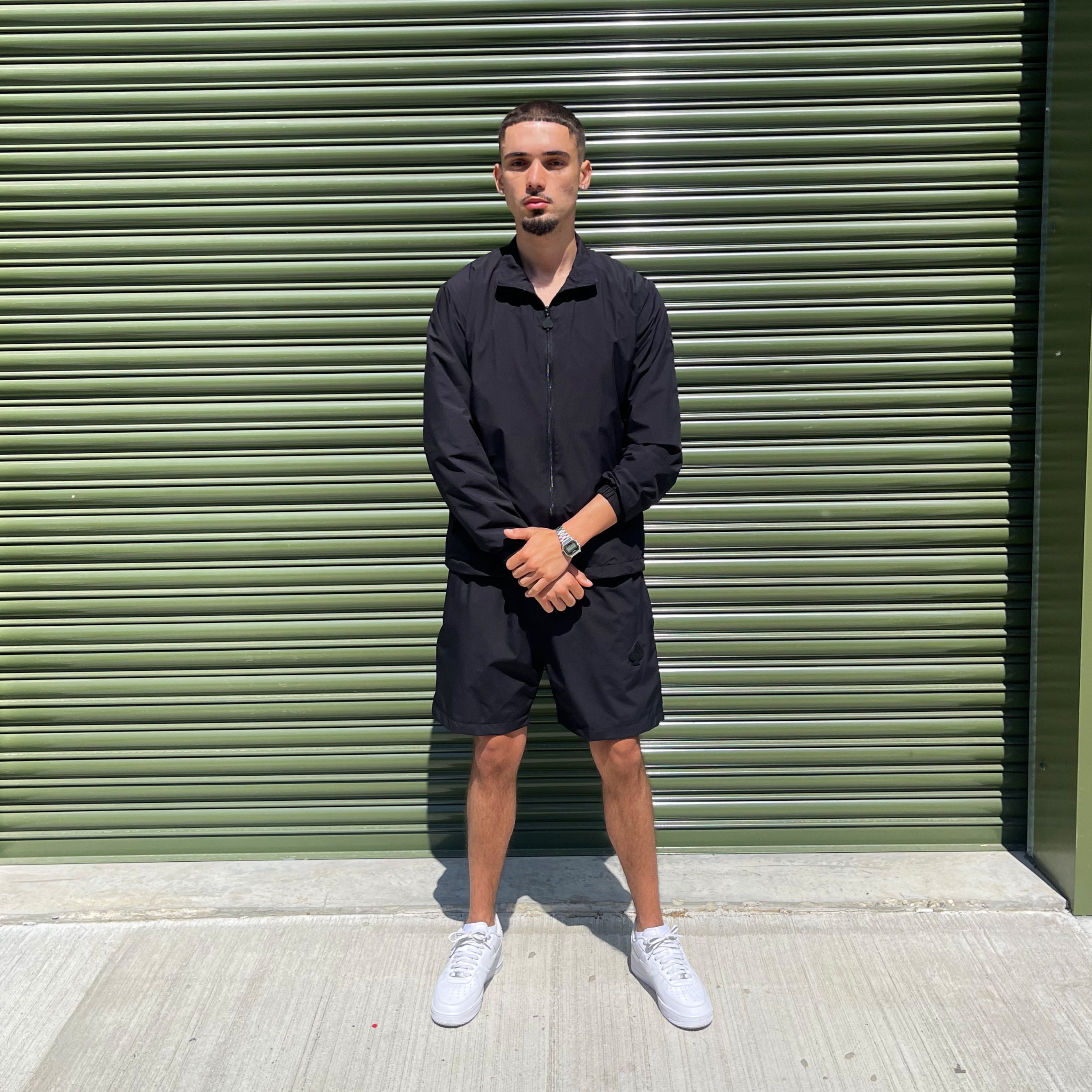 Male model wearing a black 3-piece tracksuit set from Gabos London, featuring lightweight zip jacket and matching shorts, standing against an industrial green shutter backdrop