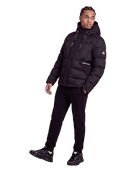 A man modeling a black hooded puffer jacket with a front zipper, zippered chest pocket, and a small white logo on the sleeve, paired with black pants and black sneakers against a plain white background.