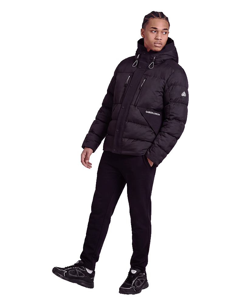 A man modeling a black hooded puffer jacket with a front zipper, zippered chest pocket, and a small white logo on the sleeve, paired with black pants and black sneakers against a plain white background.