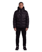 A man wearing a black hooded puffer jacket with multiple zippered pockets, black pants, and black sneakers, standing against a plain white background.