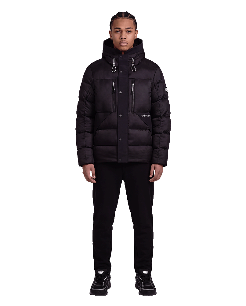 A man wearing a black hooded puffer jacket with multiple zippered pockets, black pants, and black sneakers, standing against a plain white background.