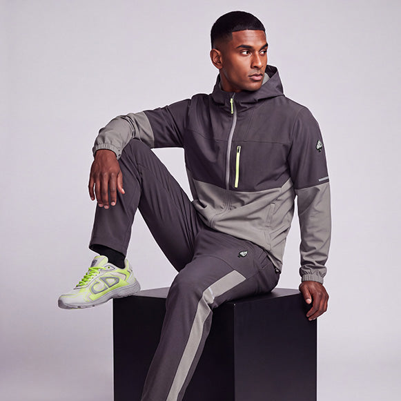 A male model wearing a dark gray and light gray color-blocked hooded jacket and matching jogger pants, styled with neon green and gray sneakers, sitting on a black cube against a light gray studio background.