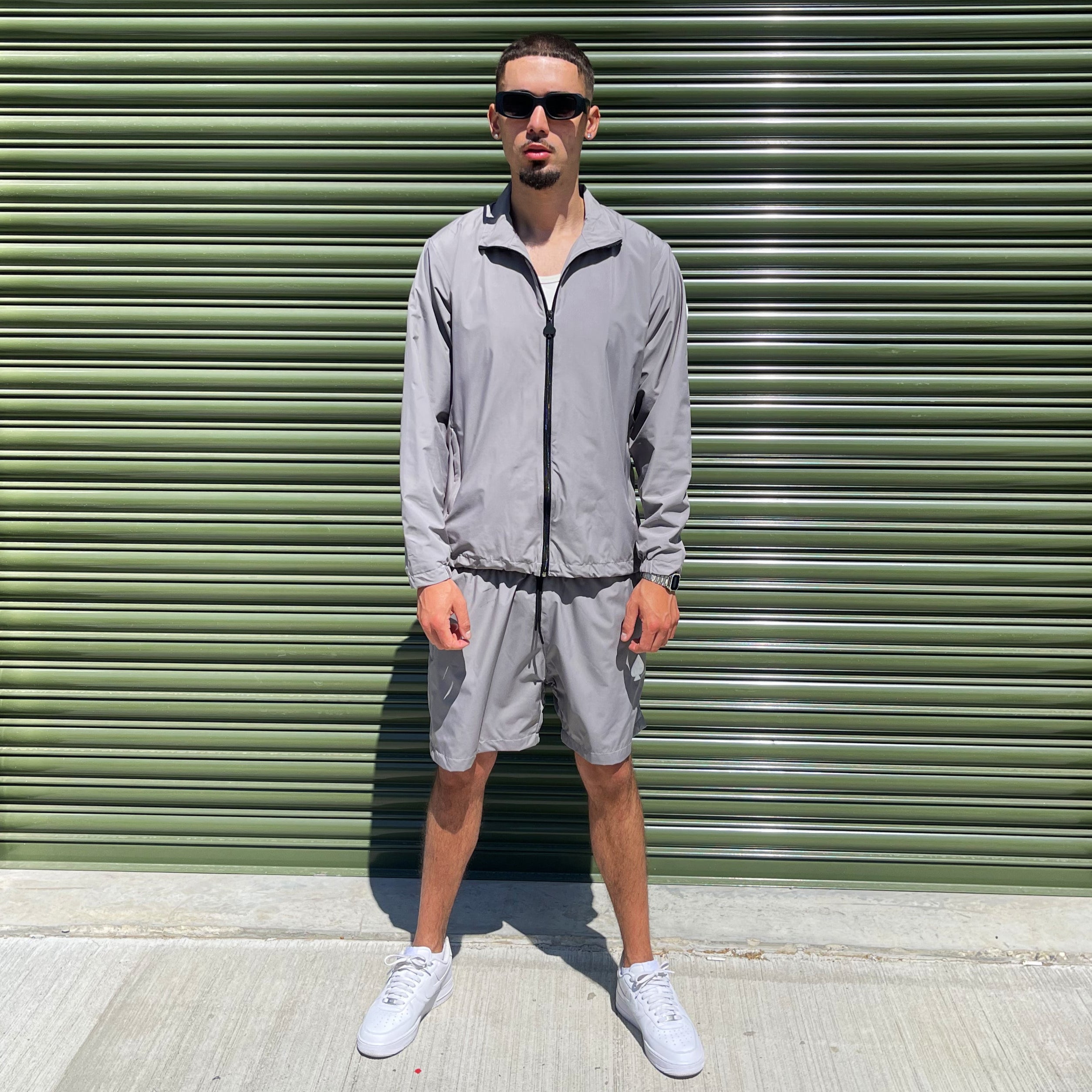 Model in grey Gabos London 3-piece tracksuit, pairing lightweight zip jacket and cargo-style shorts, seated casually in front of industrial building with sunglasses and white sneakers